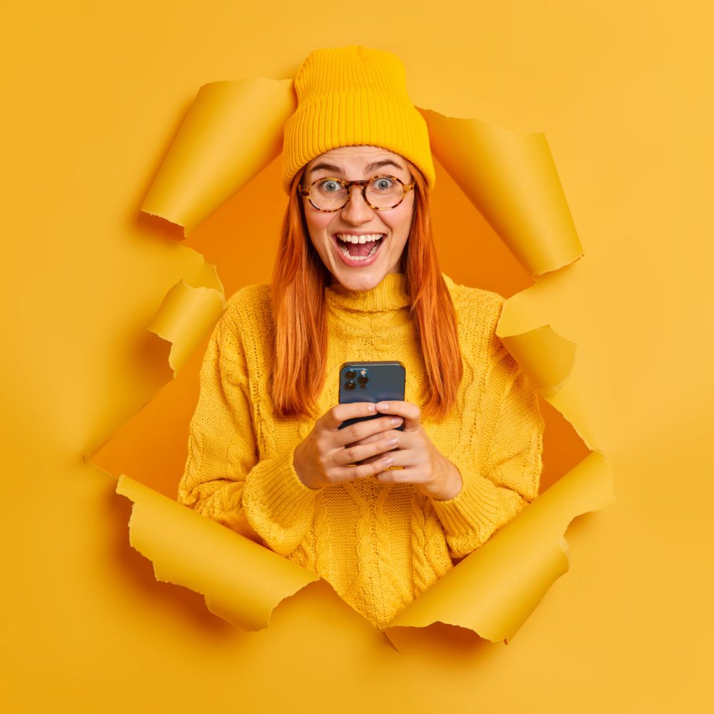 Emotional excited redhead female student uses modern mobile phone for sending text messages chats online with groupmates hears excellent news wears yellow clothes poses through hole in paper wall